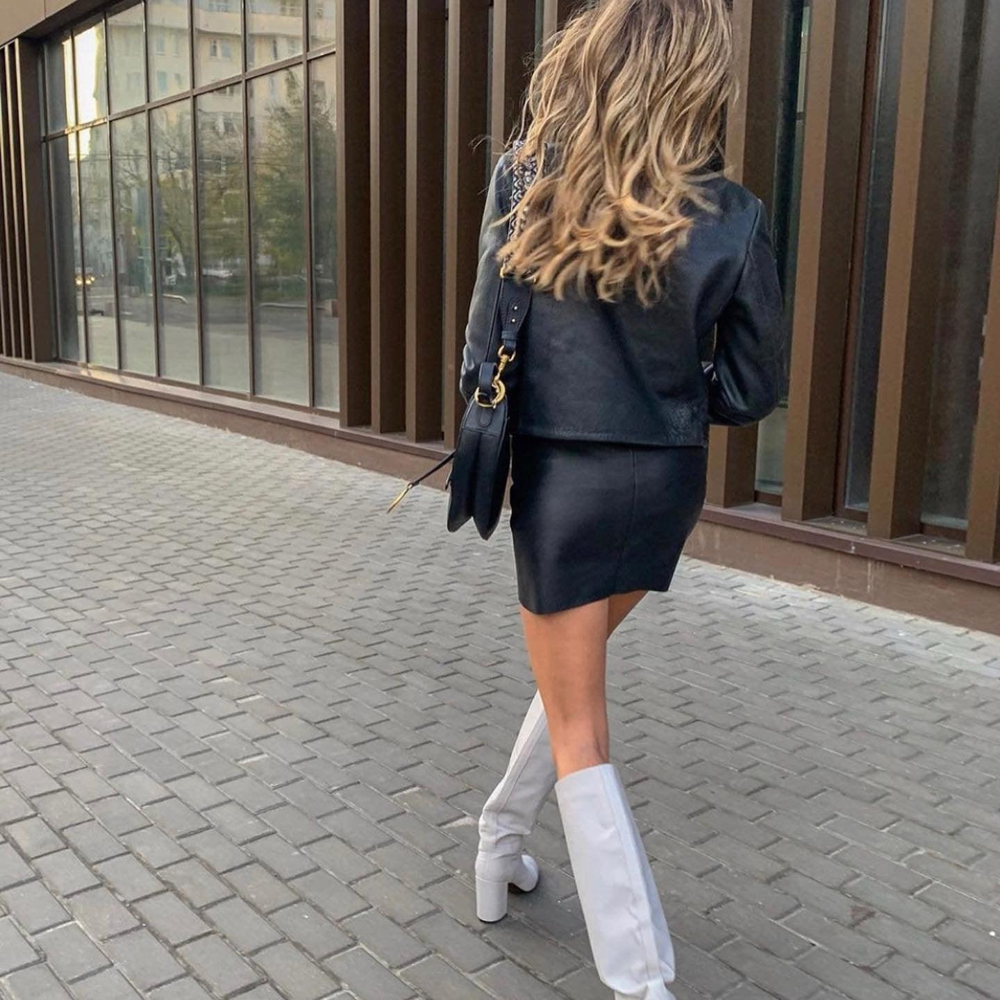 Asymmetrical black leather mini skirt styled with a fitted jacket and knee-high white boots for a chic summer look.