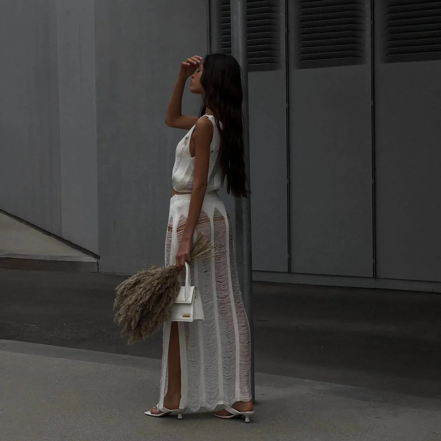 White knit two-piece set featuring a sleeveless top and a high-slit maxi skirt, styled with a chic handbag and accessories.
