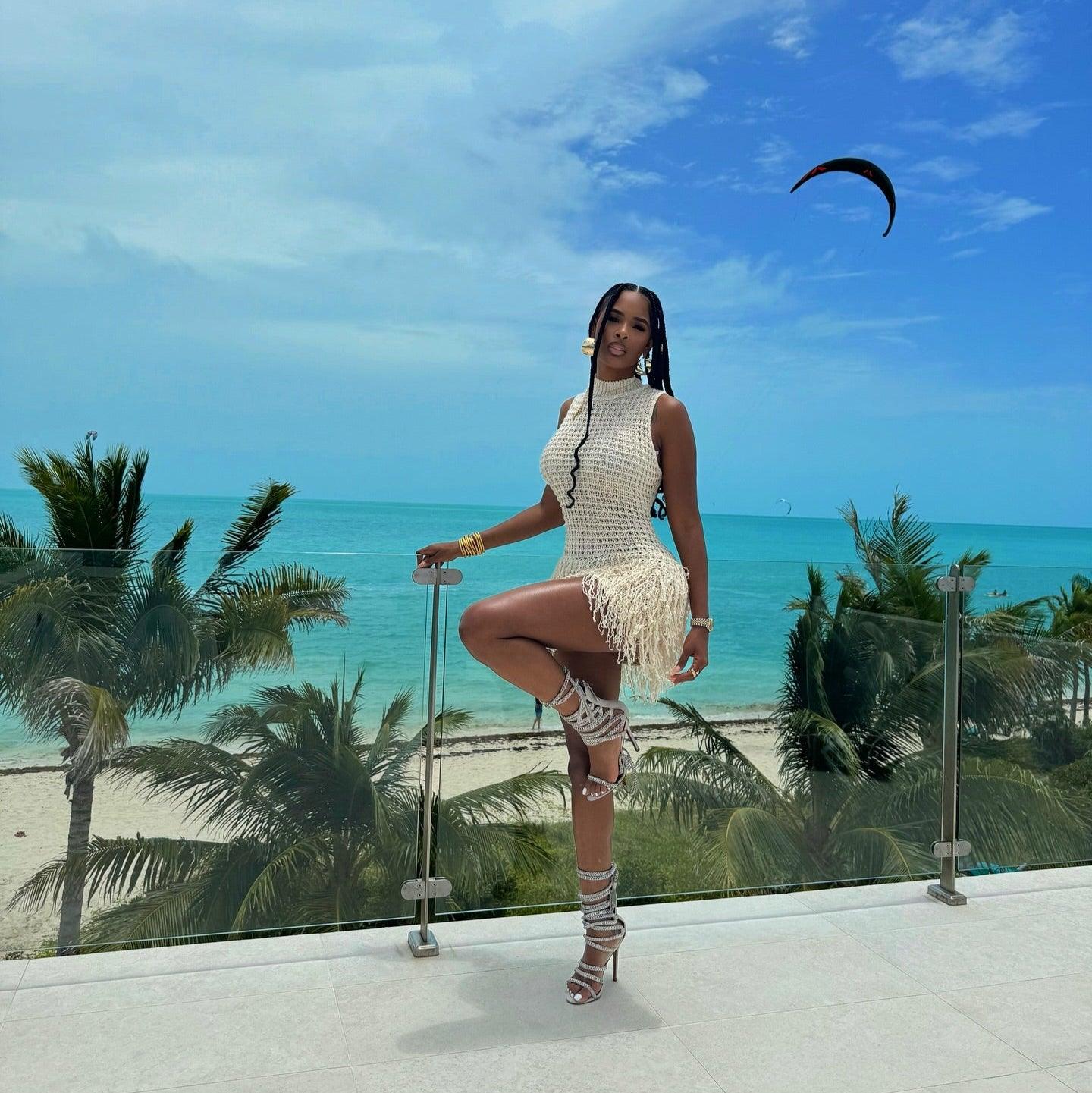 Beige knitted mini dress with fringe hemline, styled with strappy sandals against a beach backdrop.