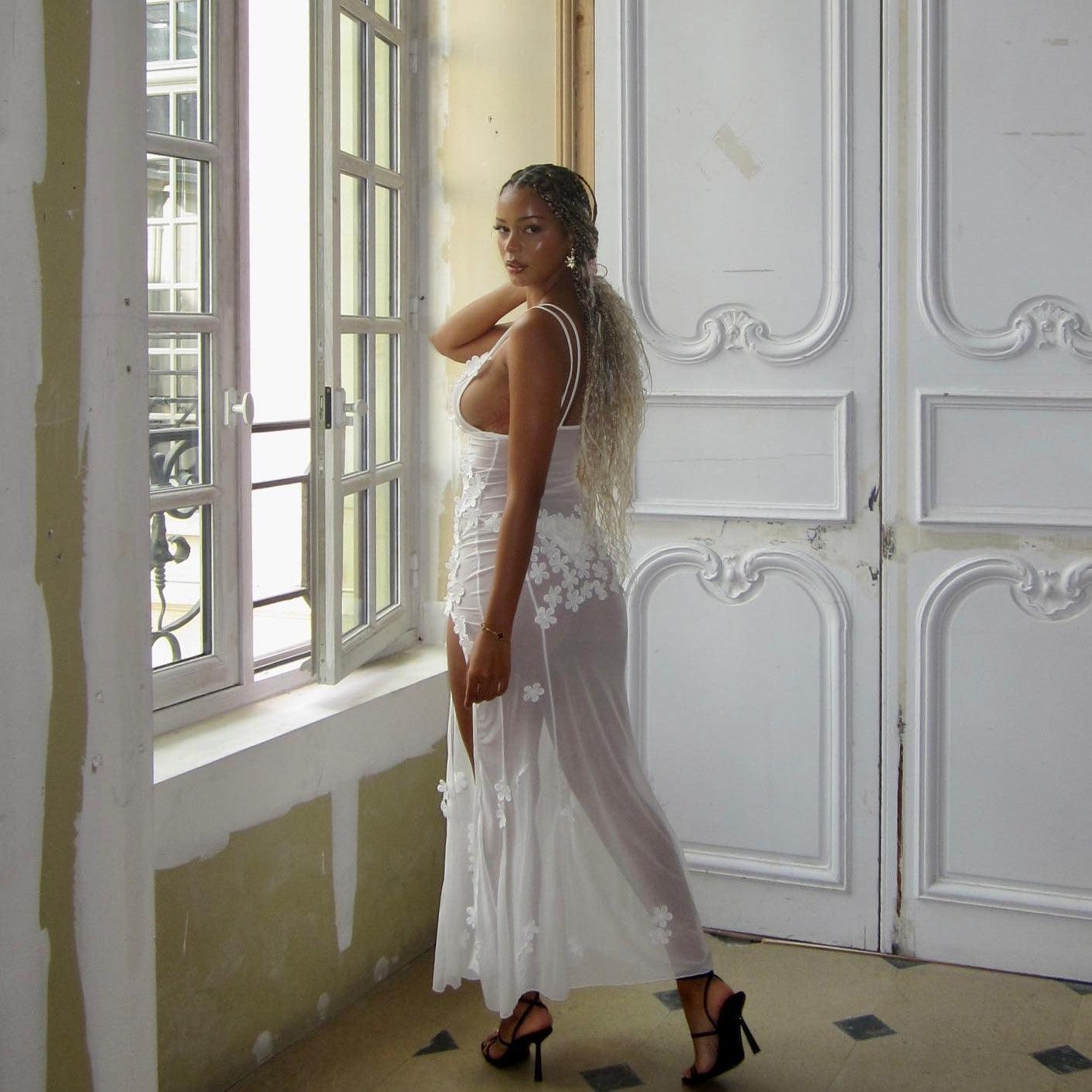 White mesh maxi dress with floral appliqués, featuring a fitted bodice and flowing skirt, styled with strappy heels.
