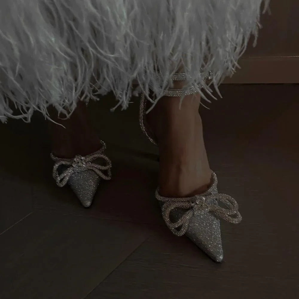 Silver glitter wrap-around bow heels with pointed toes, styled with a feathery white dress for a chic evening look.