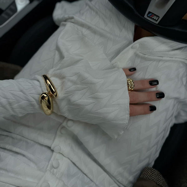 Elegant gold liquid loop bracelet worn on a hand with black nail polish, paired with a textured white garment.