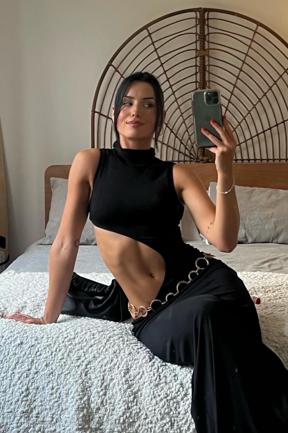 Woman taking a mirror selfie in a black outfit on a bed with a surfboard in the background.