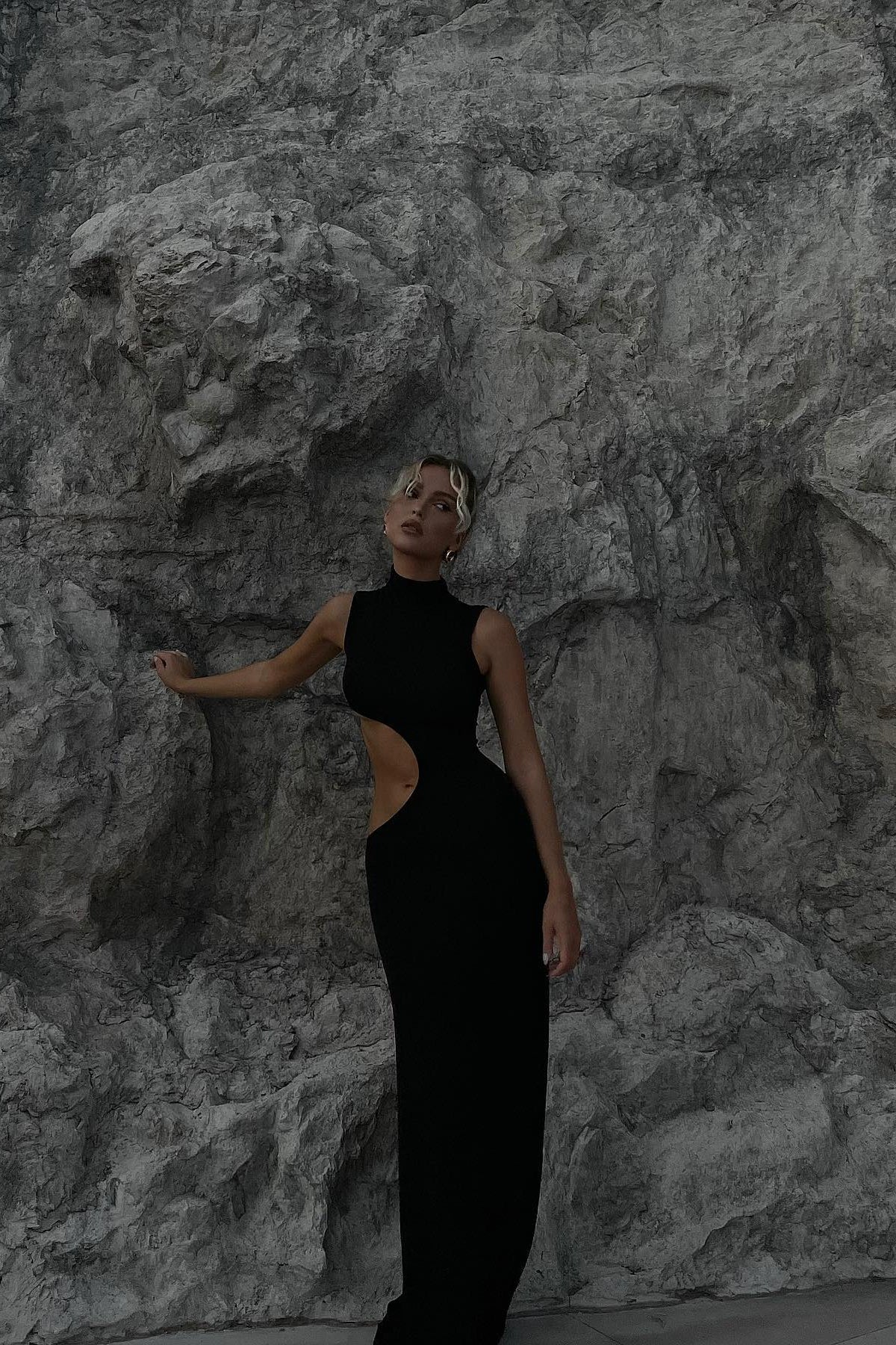 Figure-hugging black cut-out maxi dress with mock neckline against stone wall backdrop