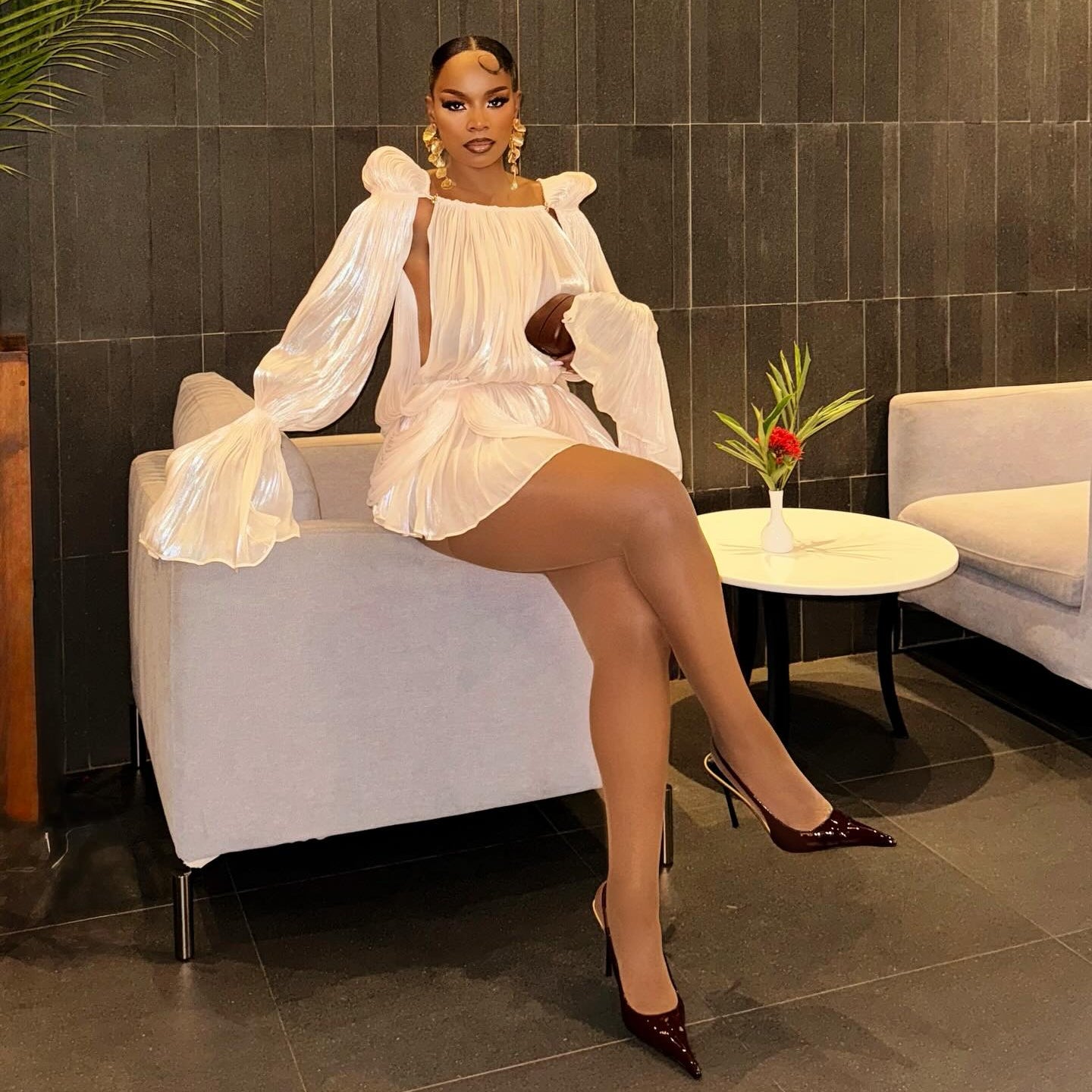 Woman in a white dress sitting on a chair in an indoor setting with a tiled wall and plant.