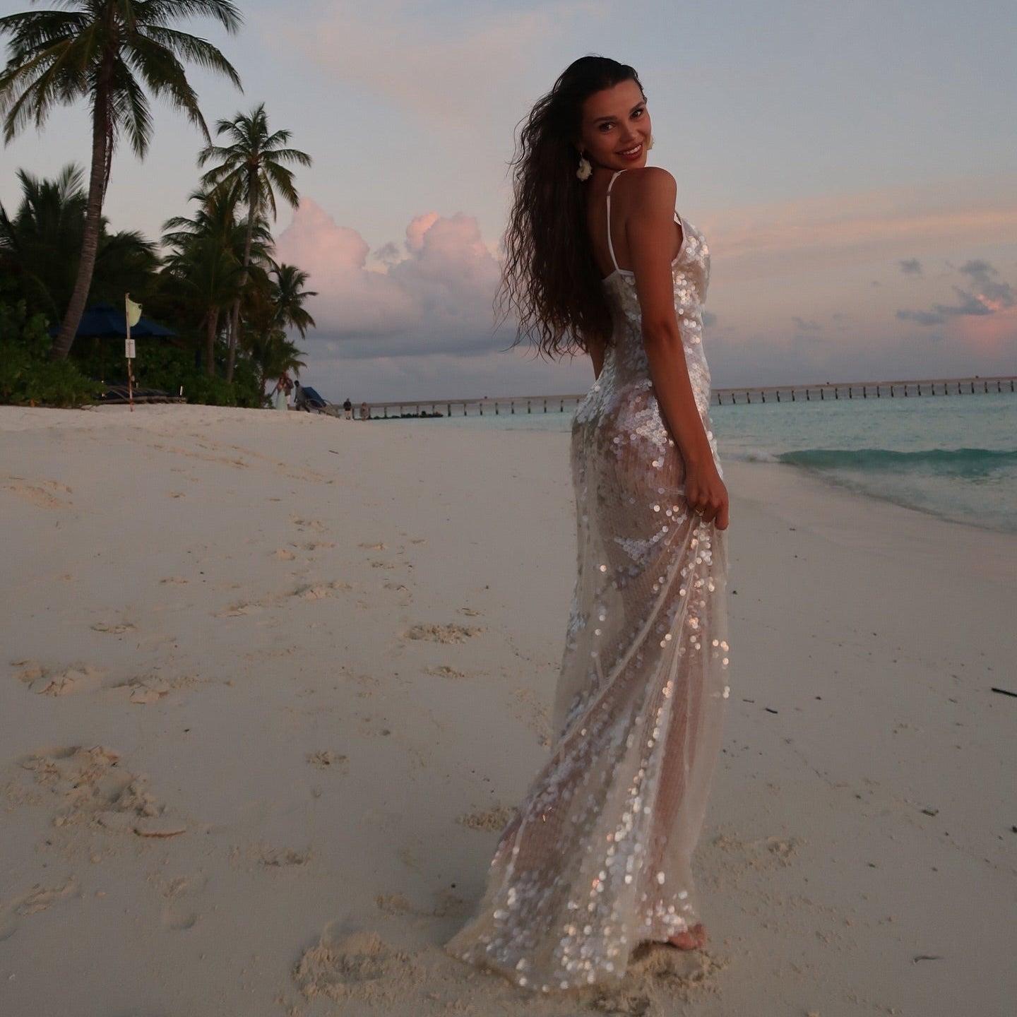 Woman in a sparkly dress standing on a beach with palm trees and a pier in the background.