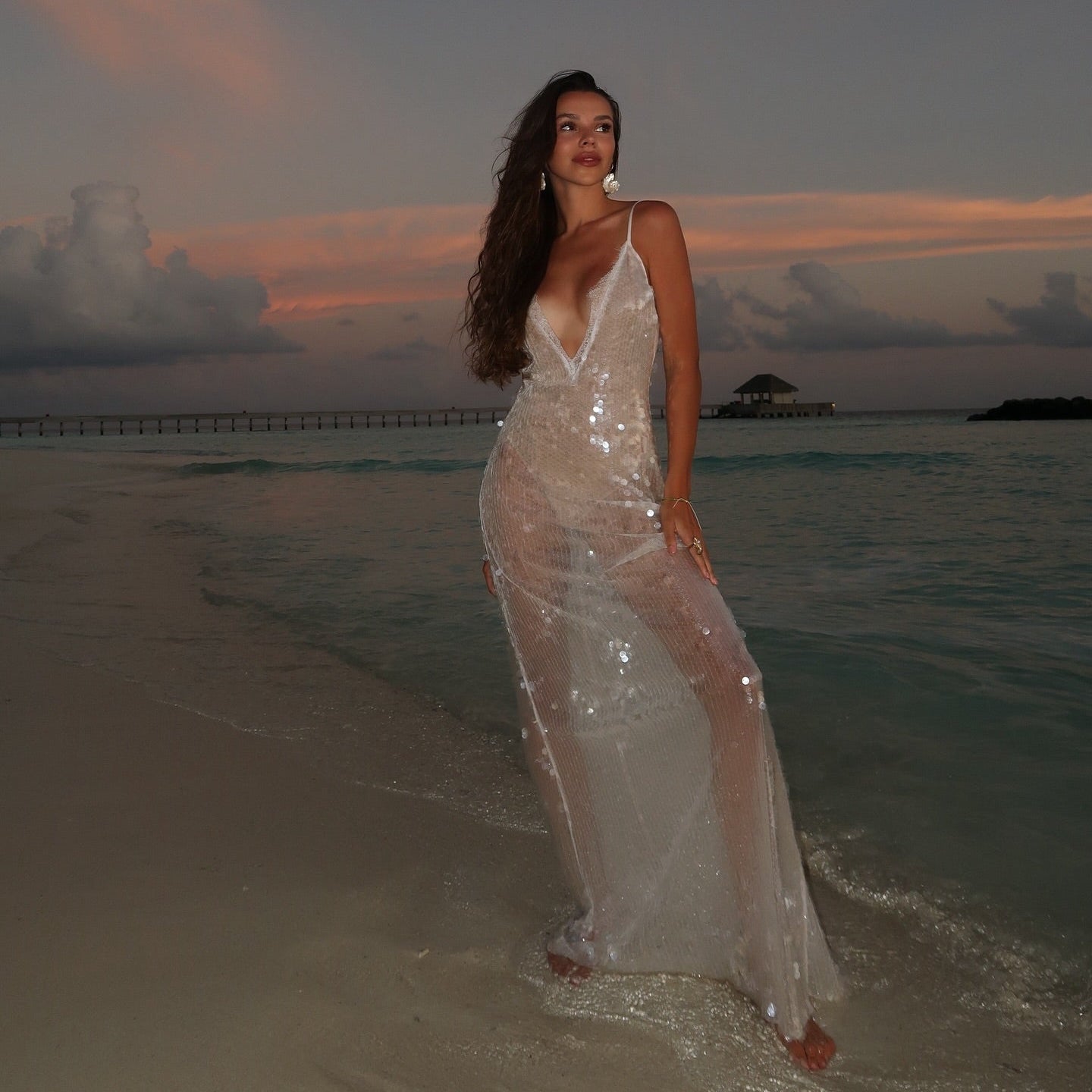 Woman in a white dress standing on a beach with a sunset and pier in the background
