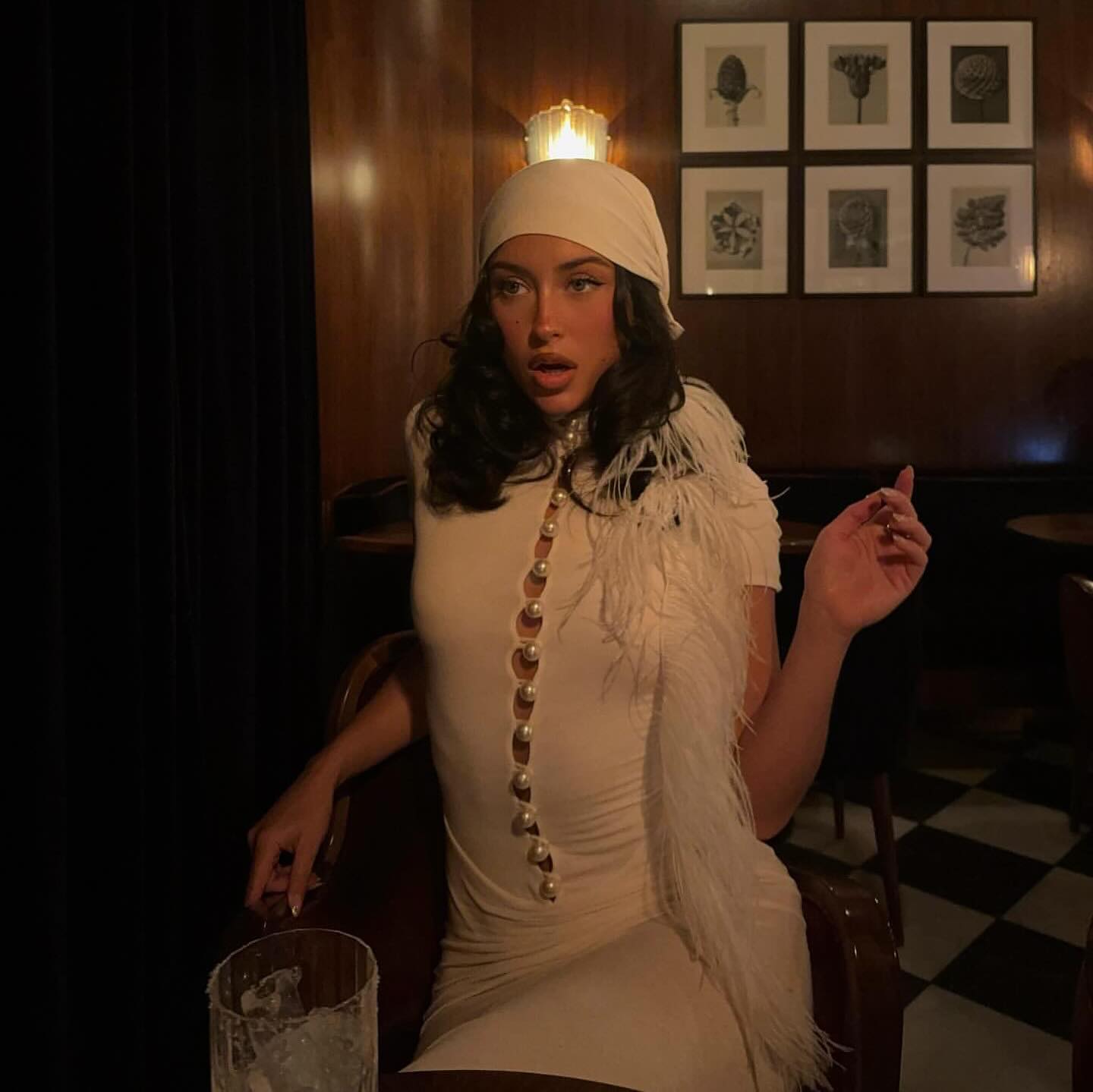 Woman in a white dress with a headscarf and fringed shawl in a dimly lit room.