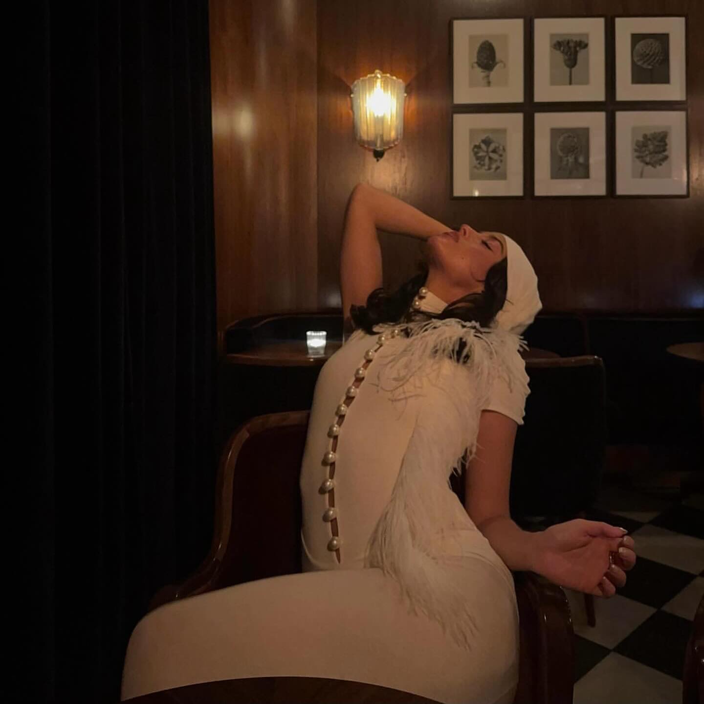 Woman in a white dress sitting in a dimly lit room with framed pictures on the wall.