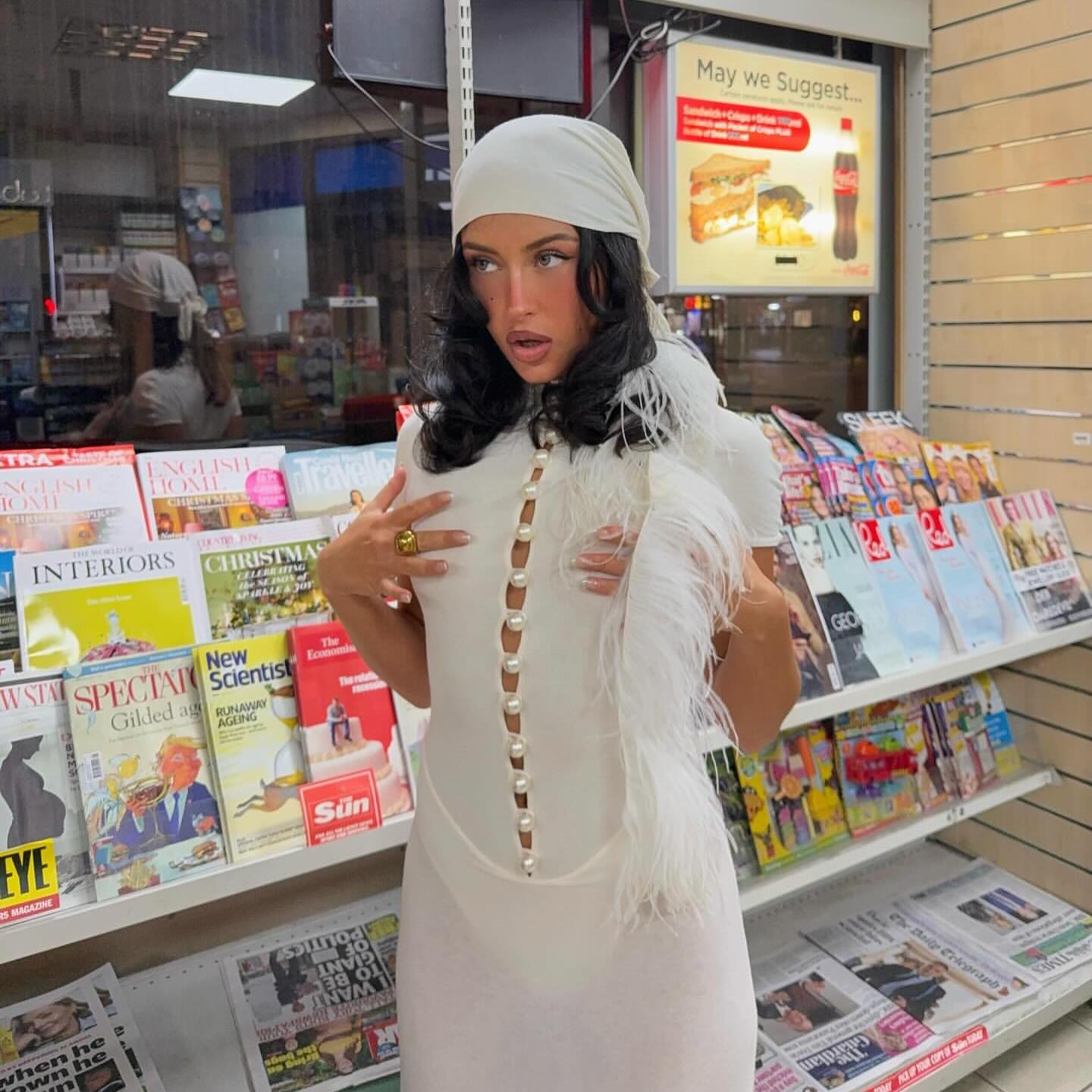 Woman in a white dress with feathers standing in front of a magazine rack.