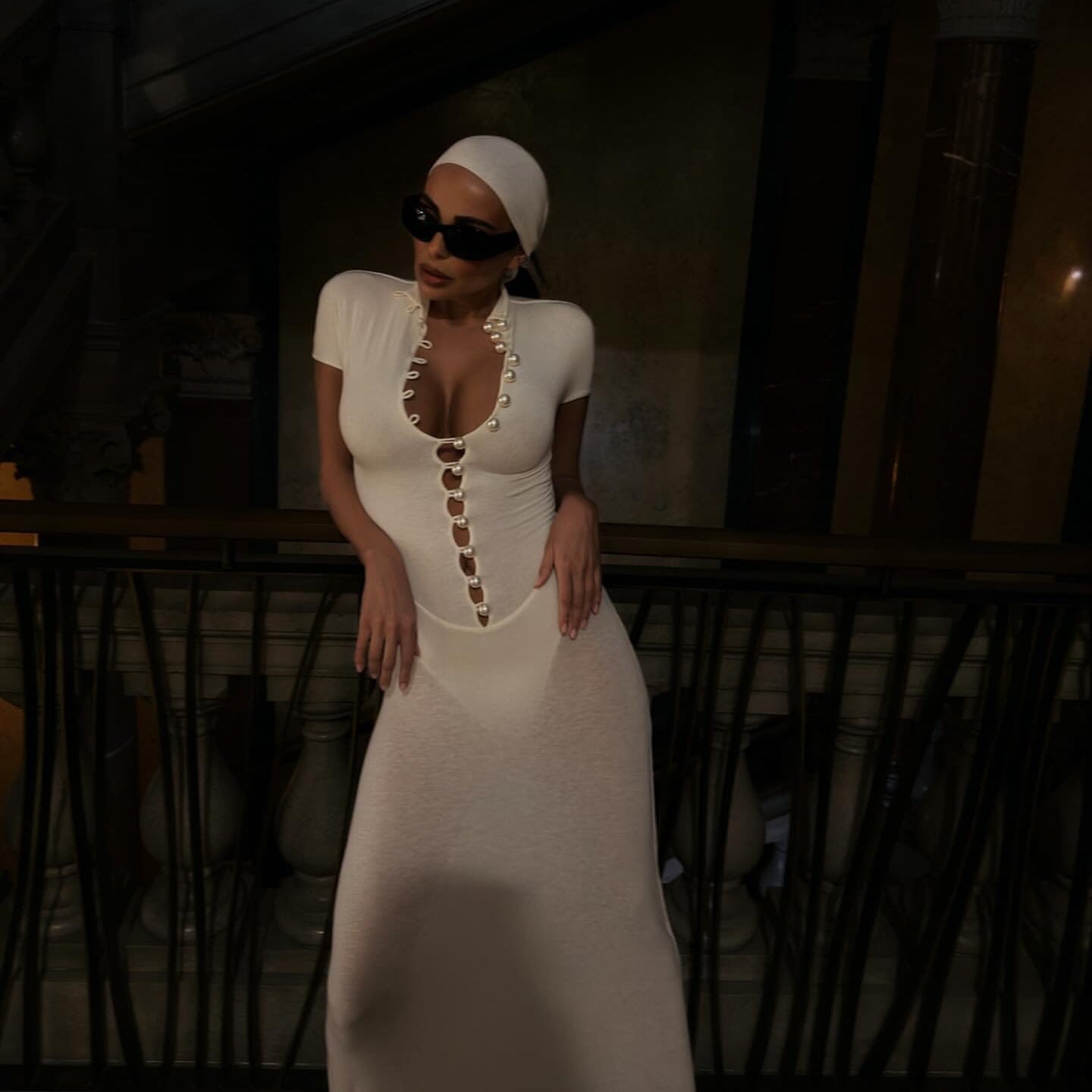 Woman in a white dress with a deep neckline and sunglasses standing in an outdoor setting.