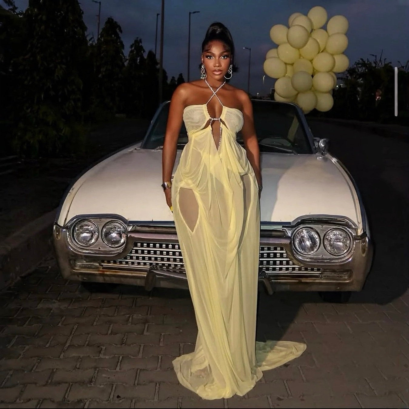 Woman in a yellow dress standing in front of an old car with balloons in the background