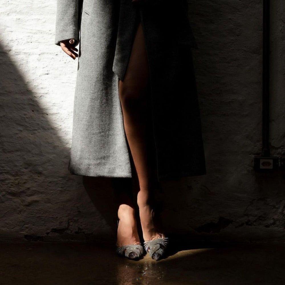 Person wearing a gray coat and shoes in a dimly lit room with a textured wall.