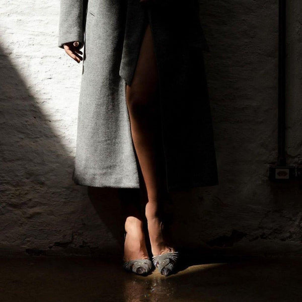 Person wearing a gray coat and shoes in a dimly lit room with a textured wall.