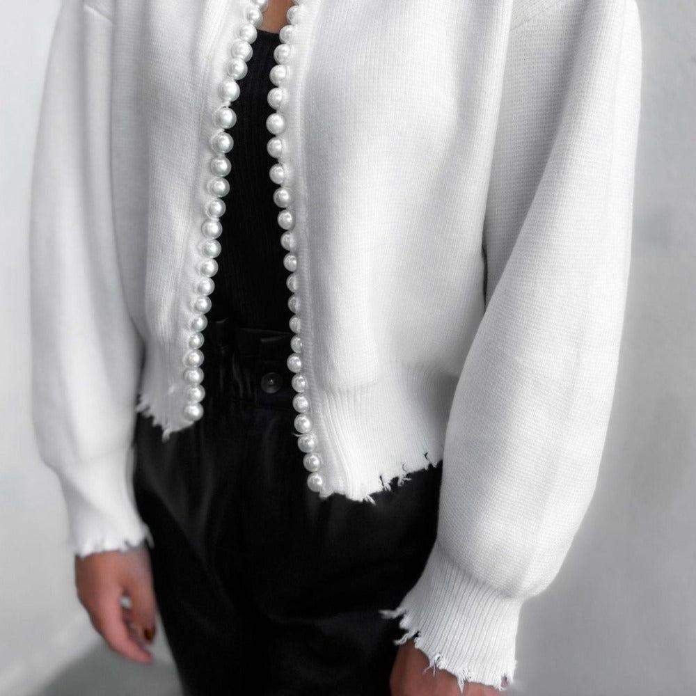 White cardigan with pearl details over a black outfit on a plain background