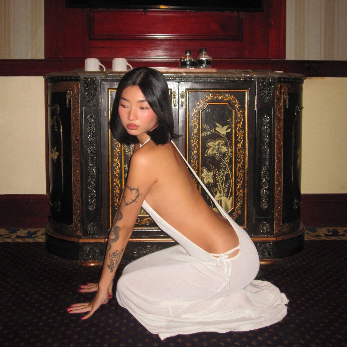 Person in a white garment kneeling in front of an ornate black cabinet.