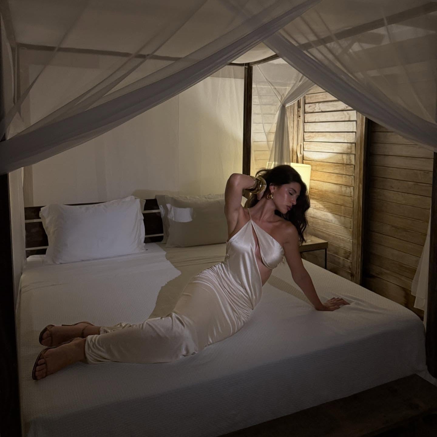 Woman in a white dress sitting on a bed in a softly lit room with wooden walls.