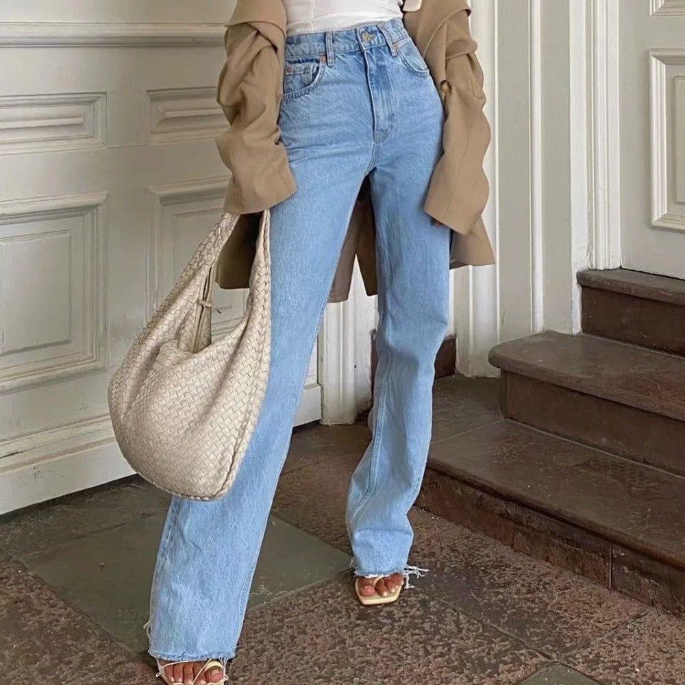 Person wearing light blue jeans and a beige coat, holding a woven handbag on a stone floor.
