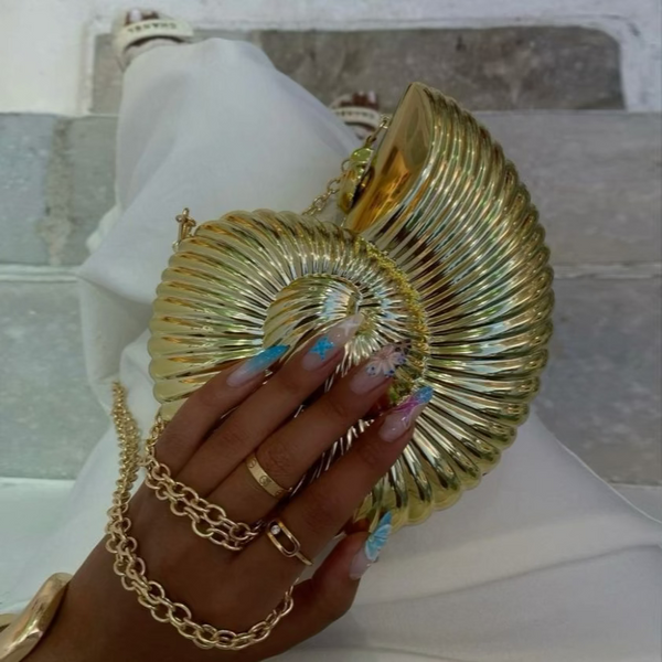 Shiny gold conch-shaped handbag with a chain strap, styled against a light outfit and colorful, manicured nails.