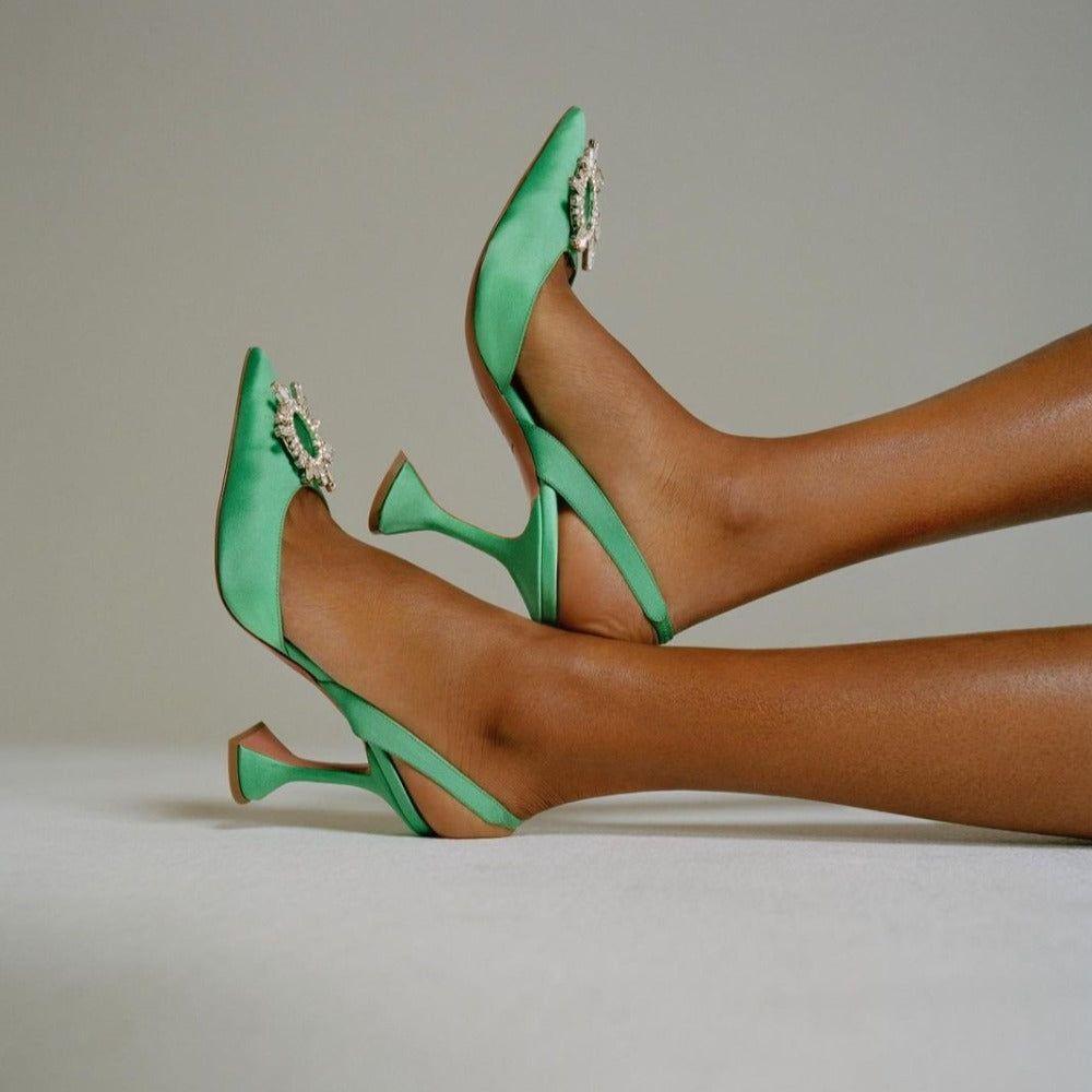 Green high-heeled shoes with embellishments worn by a person on a neutral background