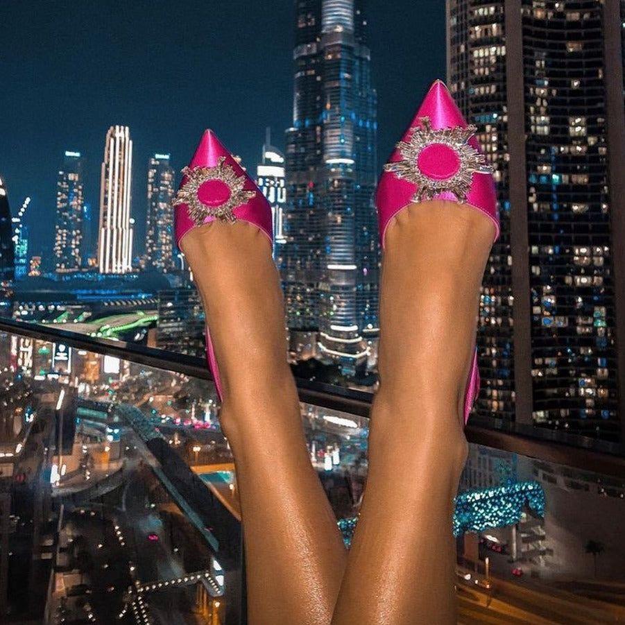 Pink high-heeled shoes with embellishments against a city skyline at night.