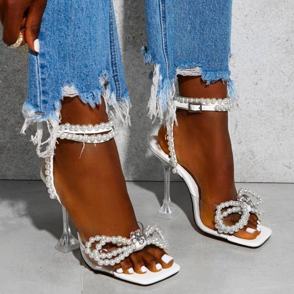High-heeled sandals with pearl embellishments worn with ripped jeans on a neutral background