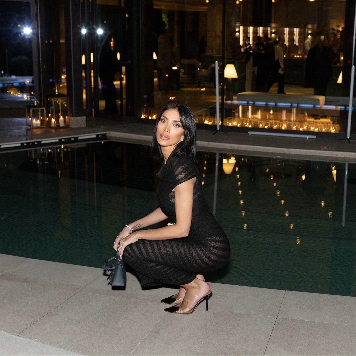 Woman in a black dress posing by a pool at night.