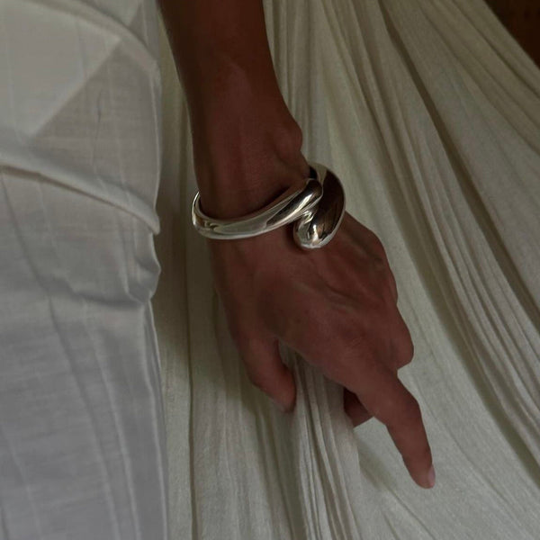 Curved silver bracelet with a sleek finish, elegantly worn on the wrist, complementing a flowing white garment.