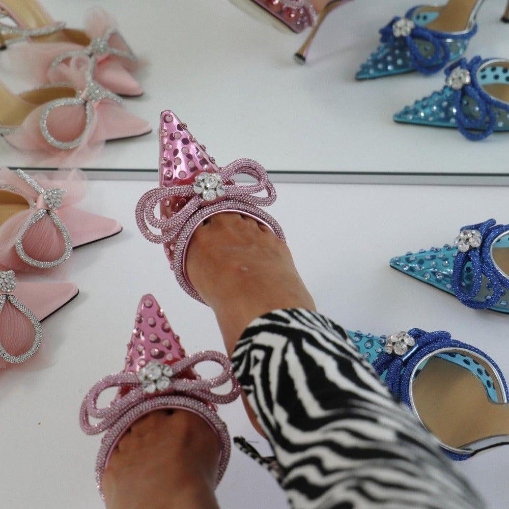 Person wearing pink high-heeled shoes with decorative bows, surrounded by other colorful shoes.