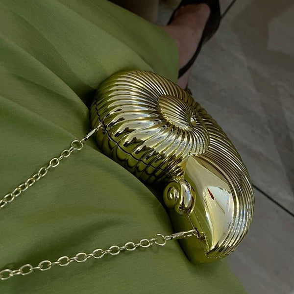 Gold conch-shaped bag with a glossy finish, styled with a chain strap, resting beside a green dress.