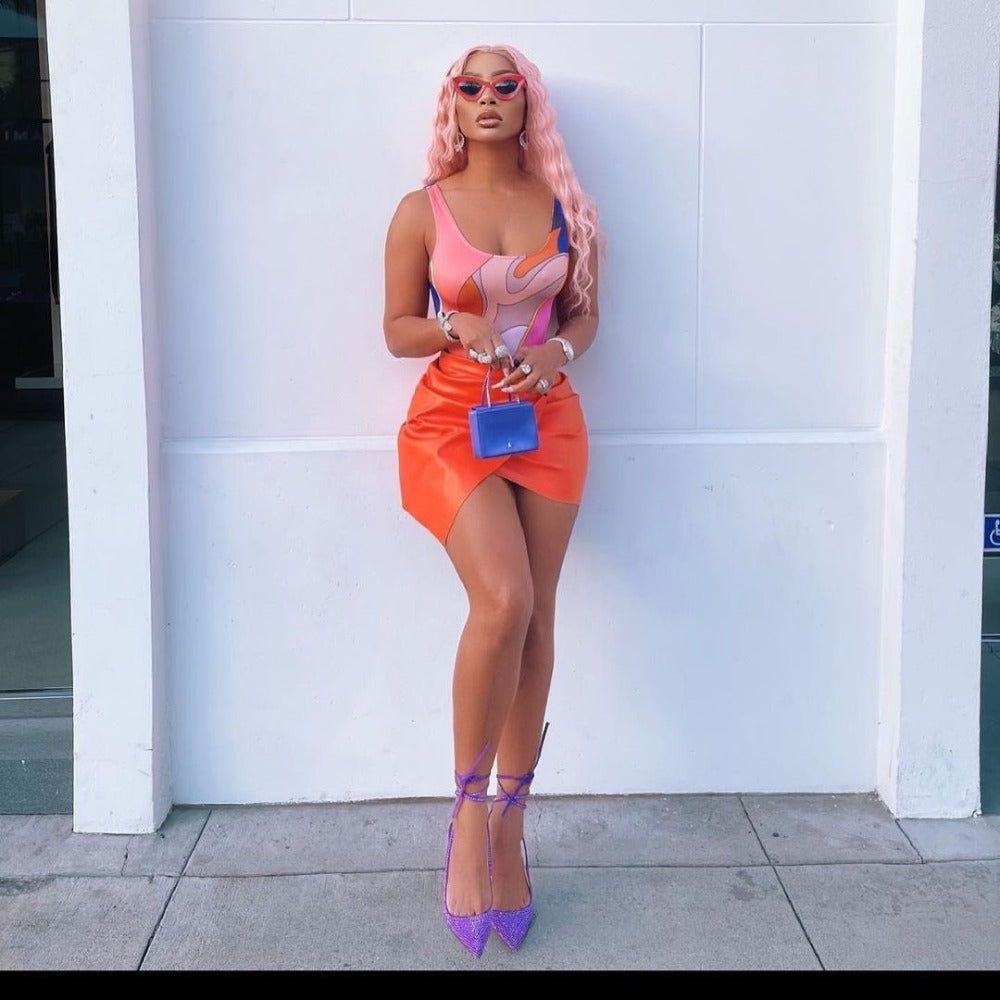 Woman in colorful outfit with pink hair and sunglasses against a white wall.