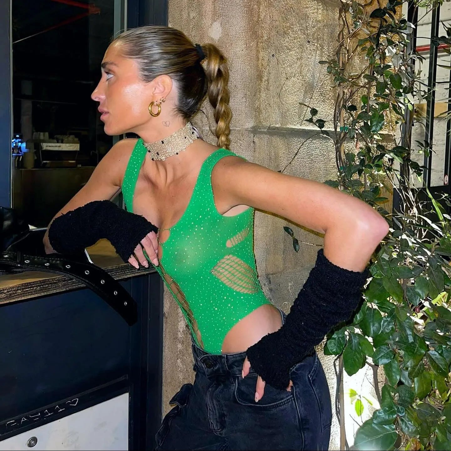 Green rhinestone embellished bodysuit with cutouts, styled with high-waisted jeans and fingerless gloves for a chic look.