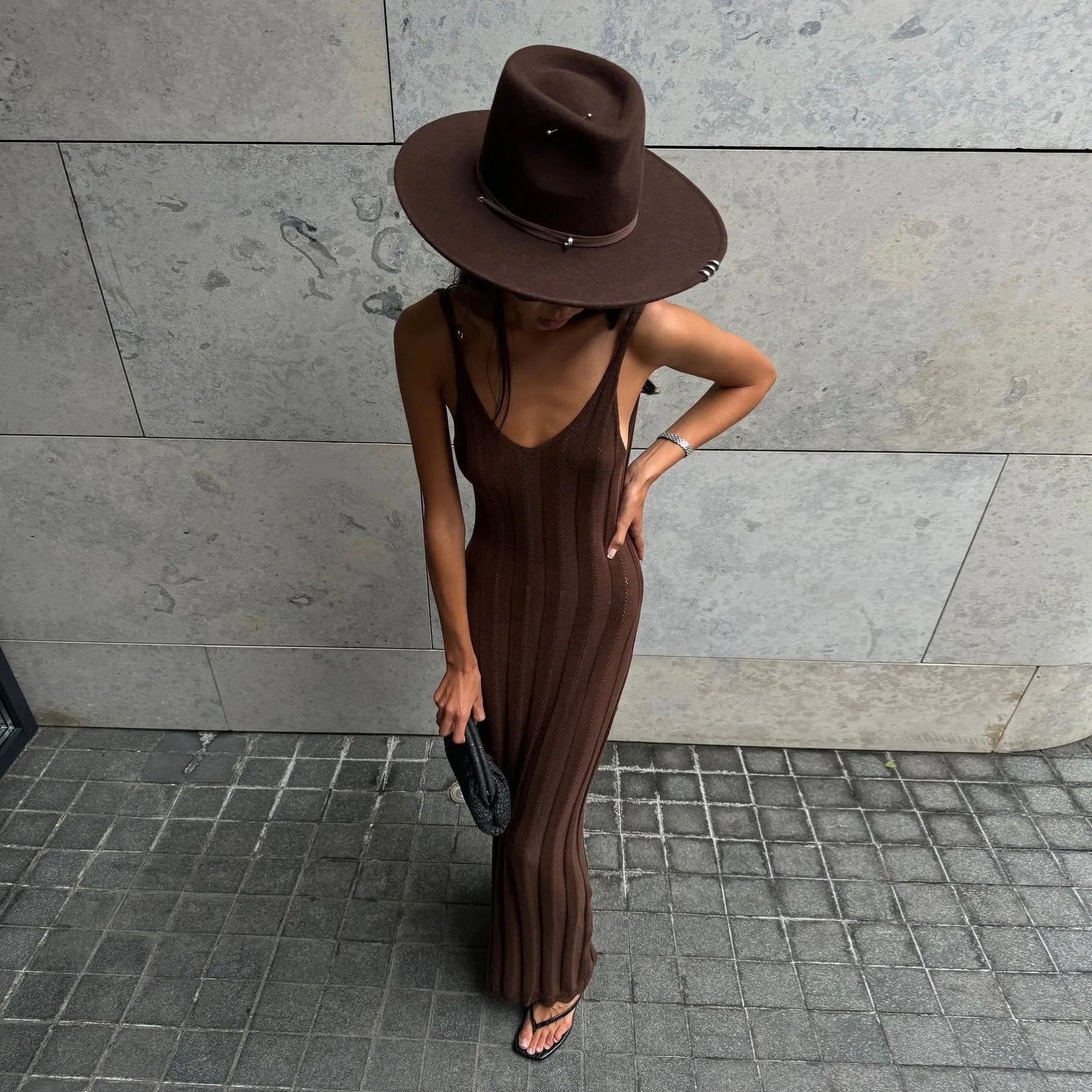 Brown knitted backless maxi dress featuring a fitted silhouette, styled with a wide-brim hat and strappy sandals.