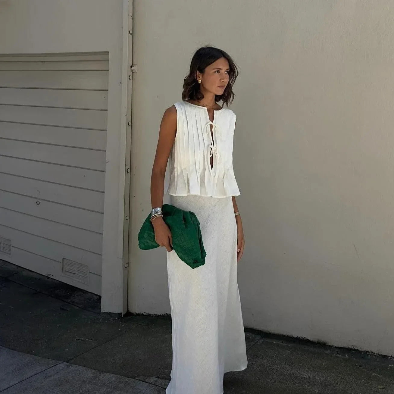 White pleated cotton top with a tie front, styled with a flowy white skirt and green clutch for a chic summer look.