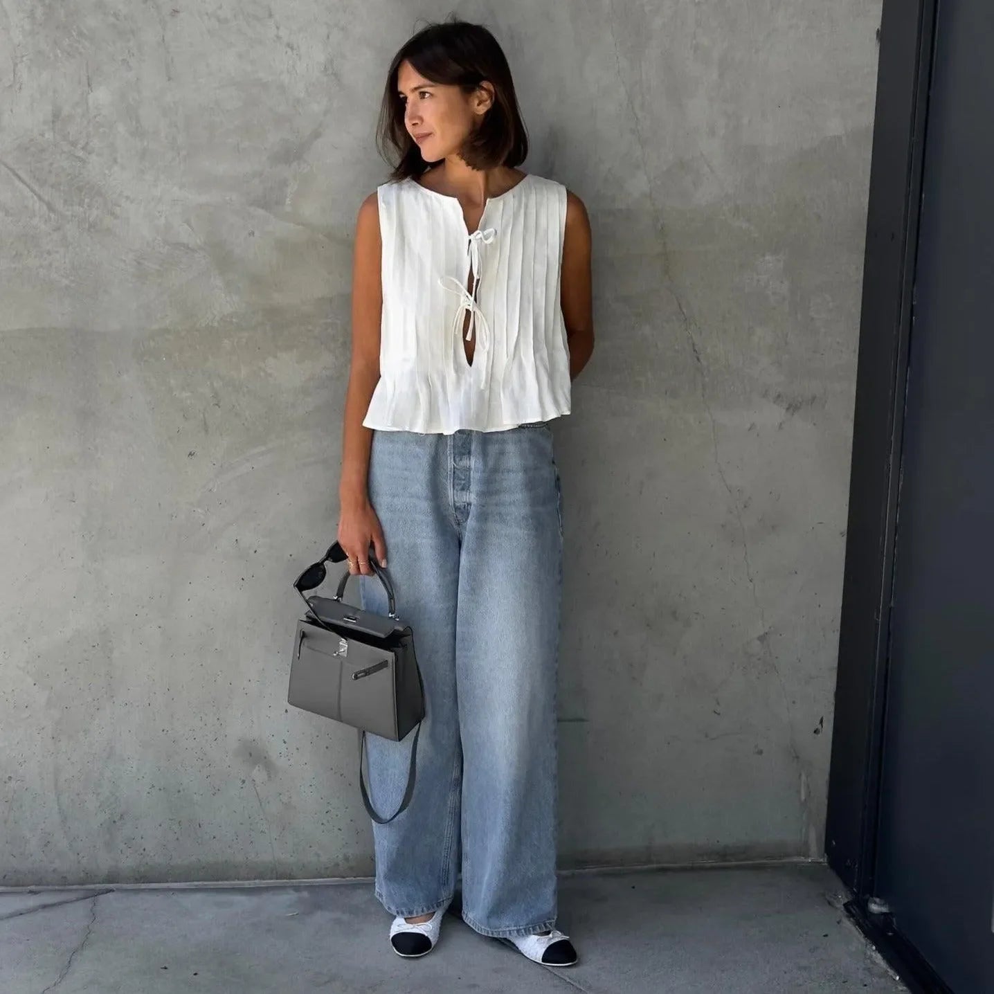 White pleated sleeveless top with a tie detail, paired with high-waisted wide-leg jeans and a chic handbag.
