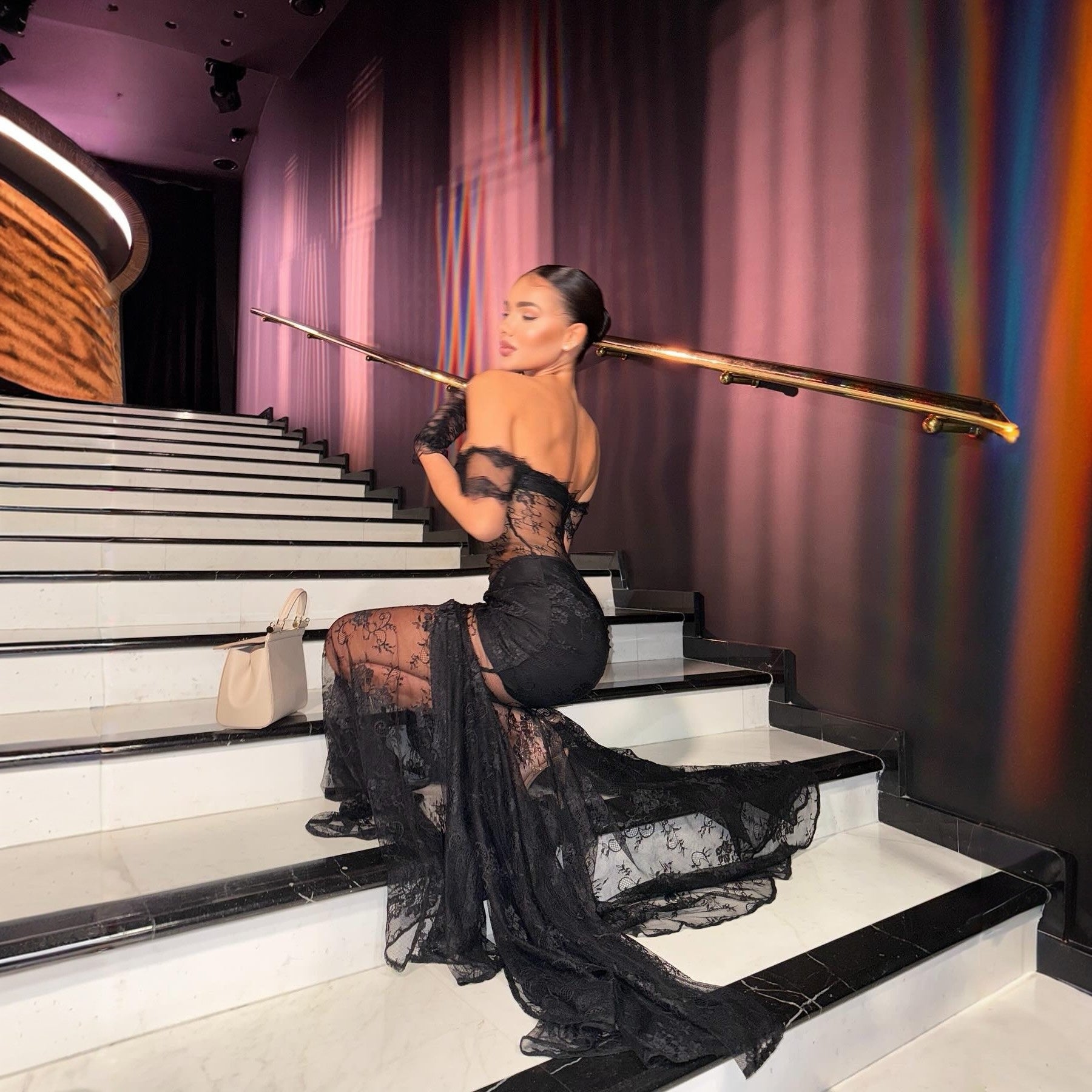 Woman in a black lace dress sitting on a staircase with colorful lighting in the background