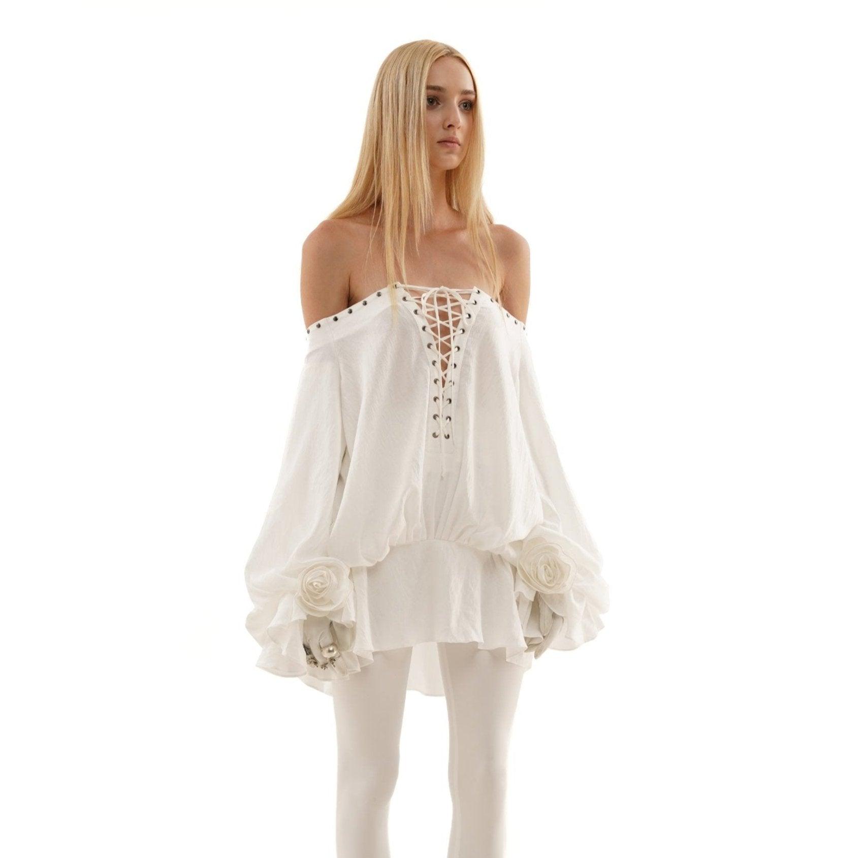 Off-shoulder white mini dress with lace-up front, flared sleeves, and floral accents, styled with fitted leggings.