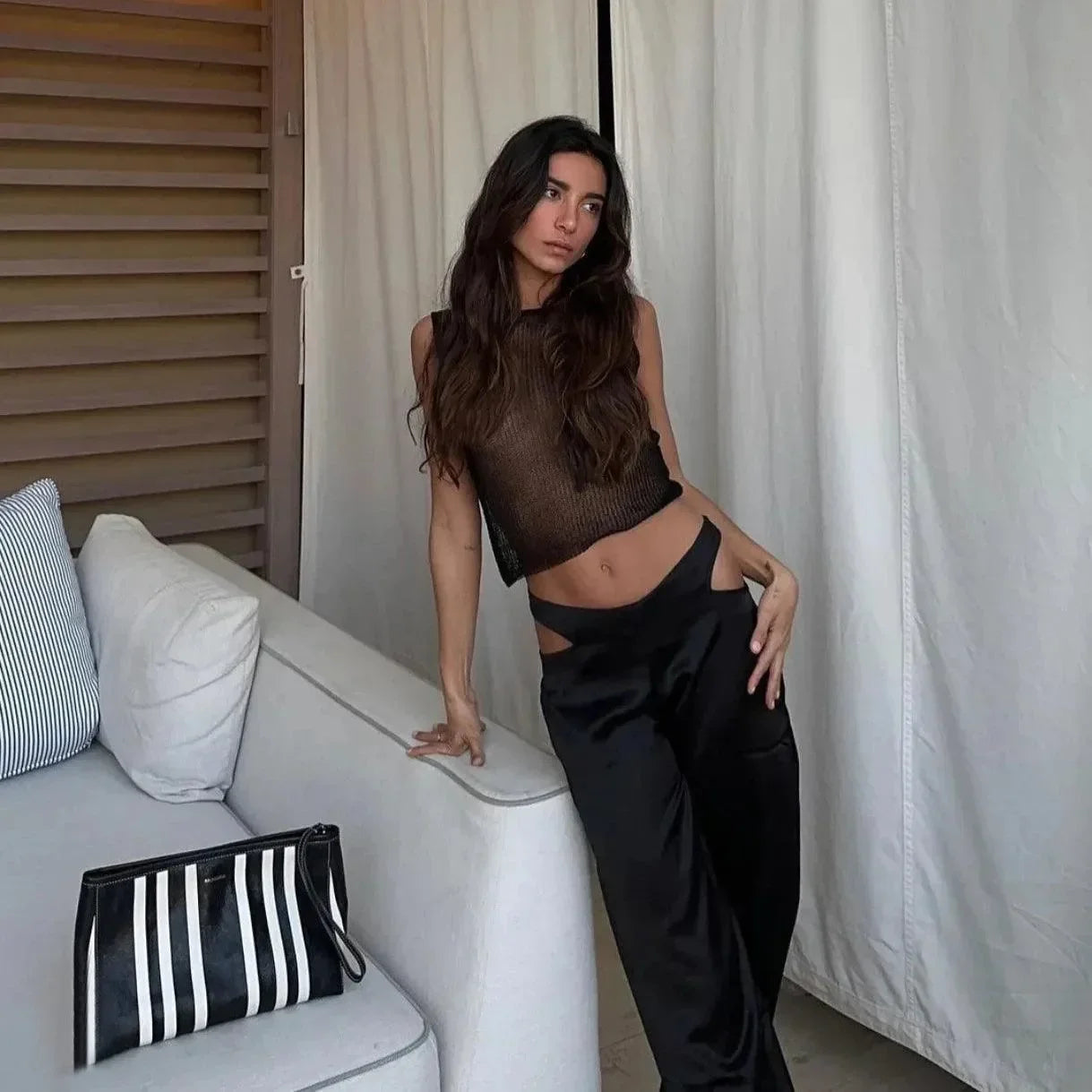 Sheer black sleeveless top with a high neck, styled with loose black pants and a striped handbag for a chic look.