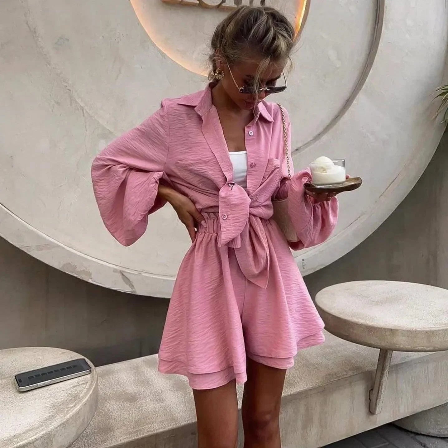 Light pink, two-piece set featuring a tied shirt with bell sleeves and matching shorts, styled with a casual tank top.