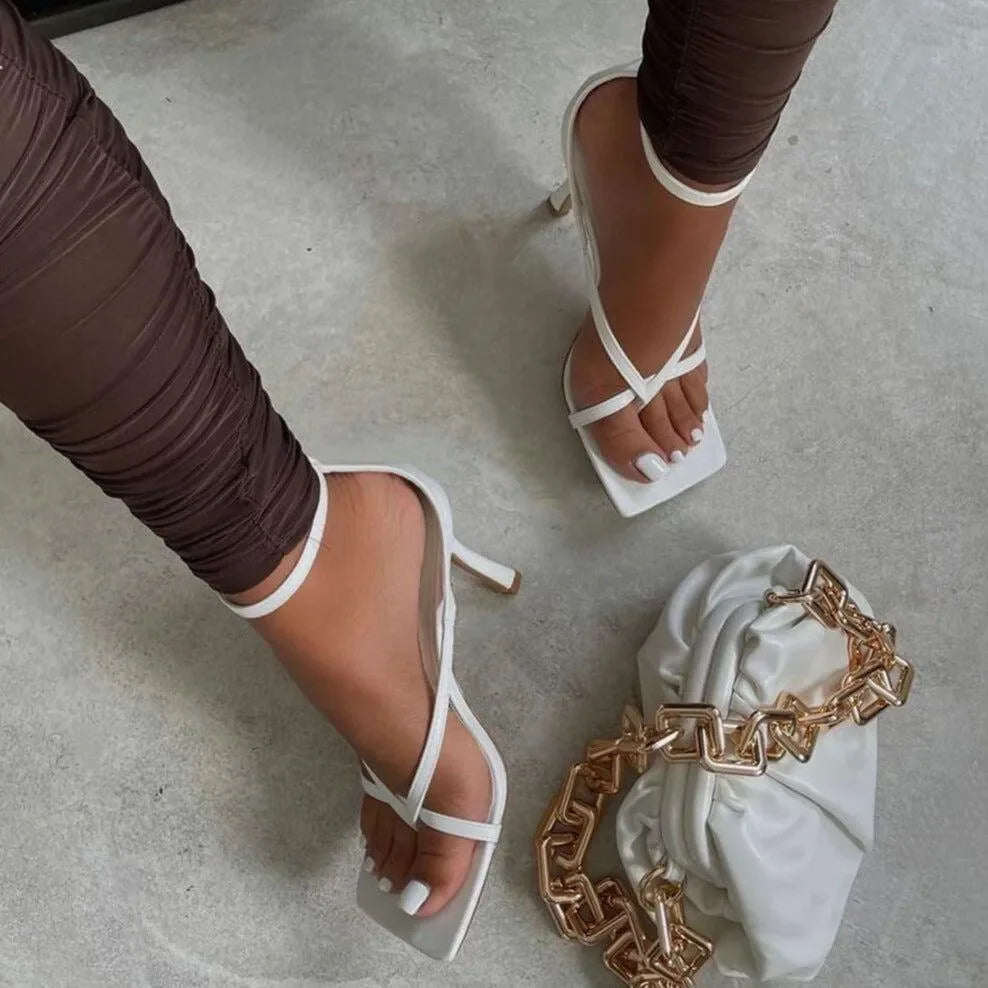 White high-heeled sandals worn by a person on a light gray floor with a gold chain and white fabric.